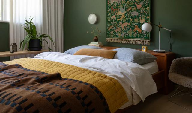 Made bed with mustard quilt and brown blanket in a green-walled bedroom with tapestry, plant and bedside lamp