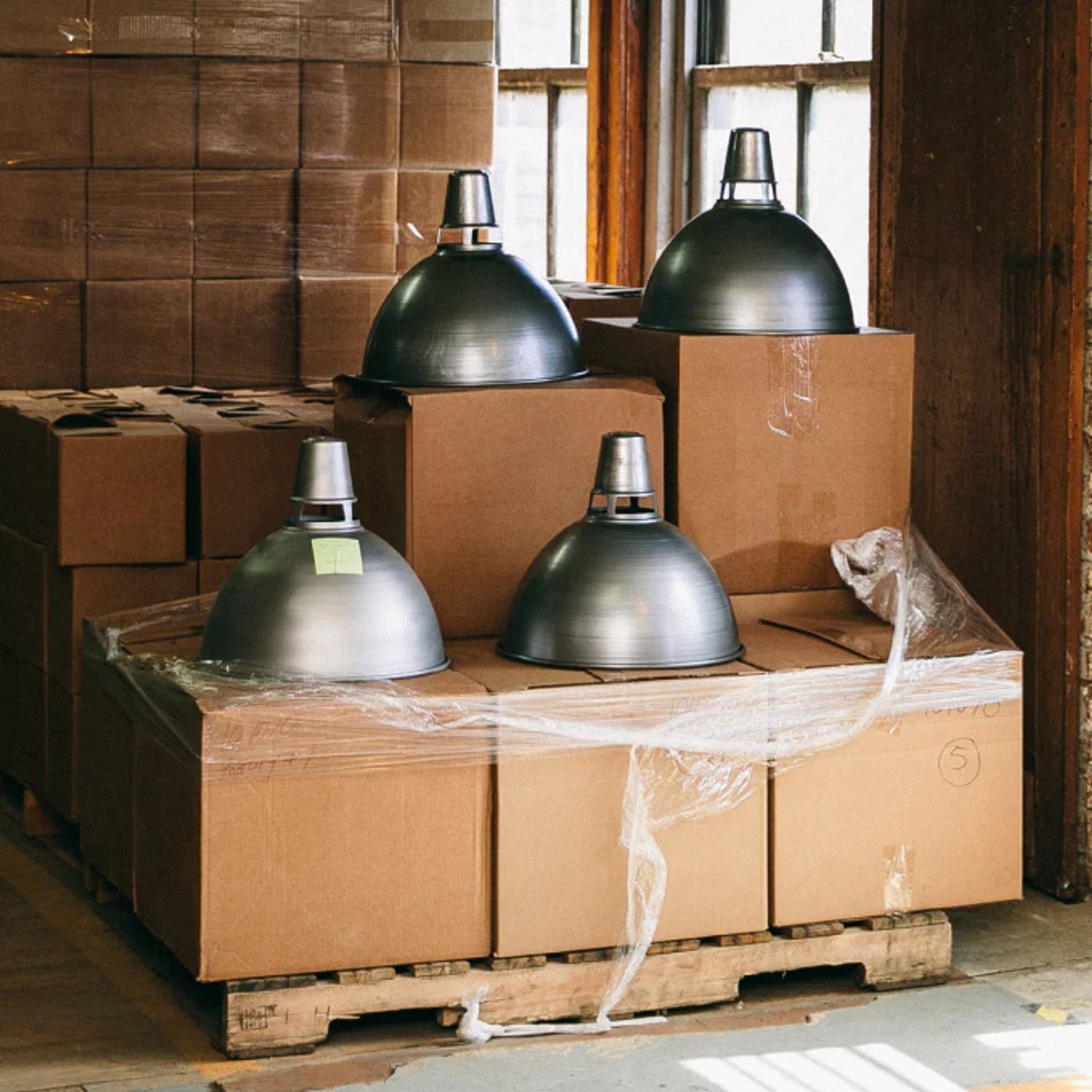 Four metal pendant lamp shades resting on stacked cardboard boxes on a wooden pallet in a sunlit warehouse