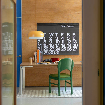 Dining table styled as a workspace with a green chair, yellow pendant lamp, books and coffee cup, and a large wall calendar reading “2024 October”