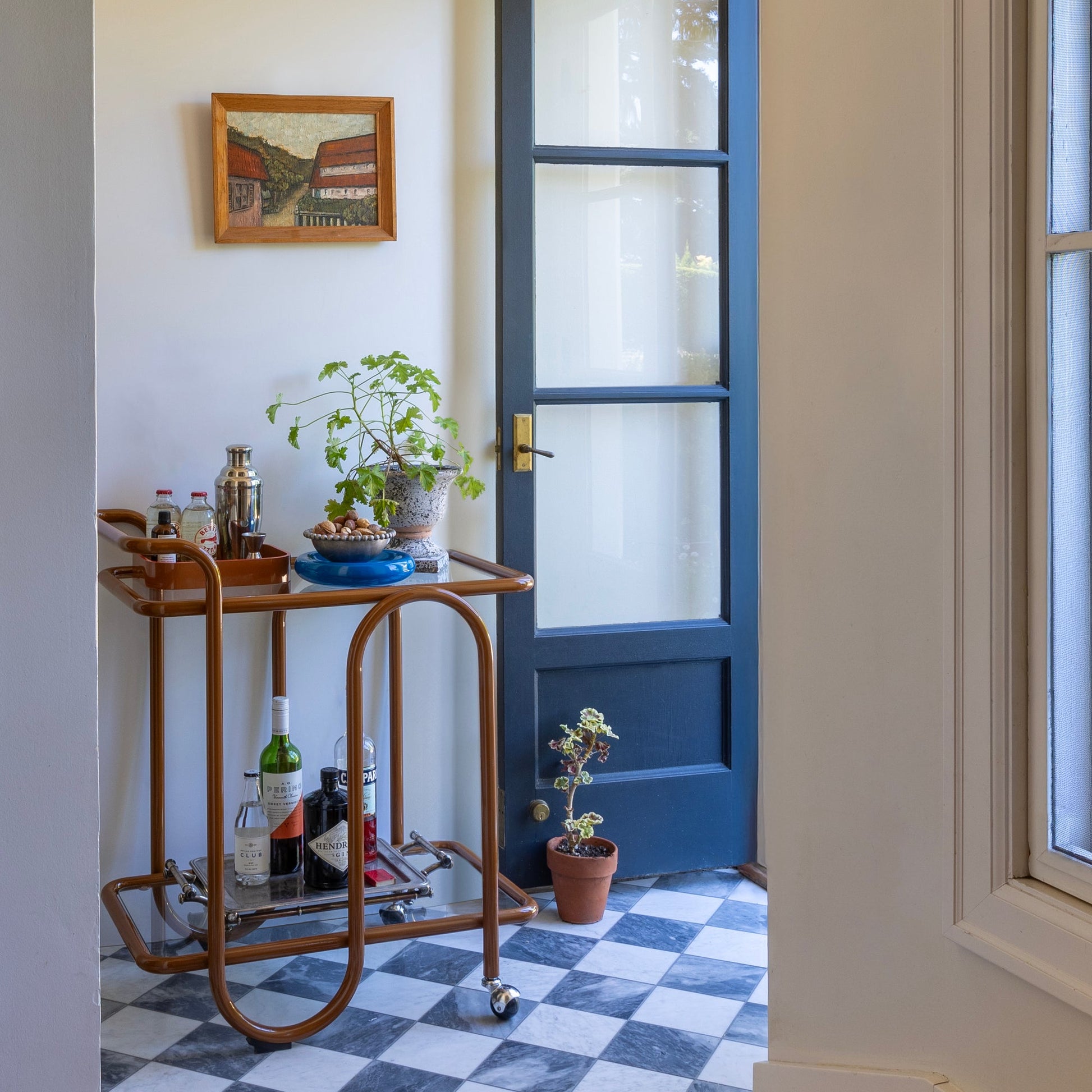 Angled view of Lolly Bar Cart with bent-steel frame, glass shelves, and ball-caster wheels beside a blue glass-panel door