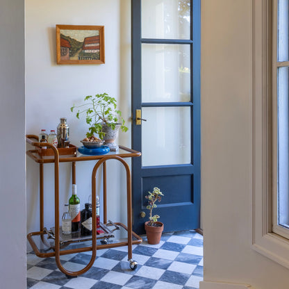 Angled view of Lolly Bar Cart with bent-steel frame, glass shelves, and ball-caster wheels beside a blue glass-panel door