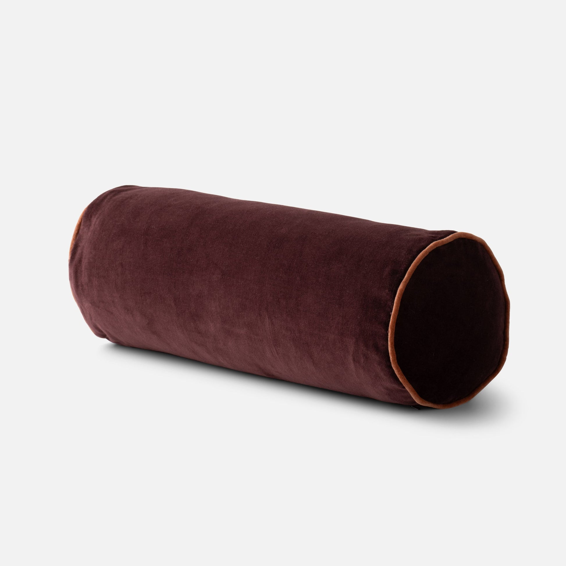 Angled close up of Velvet Piped Pillow cylinder showing raisin velvet with contrast rust piping on light gray background