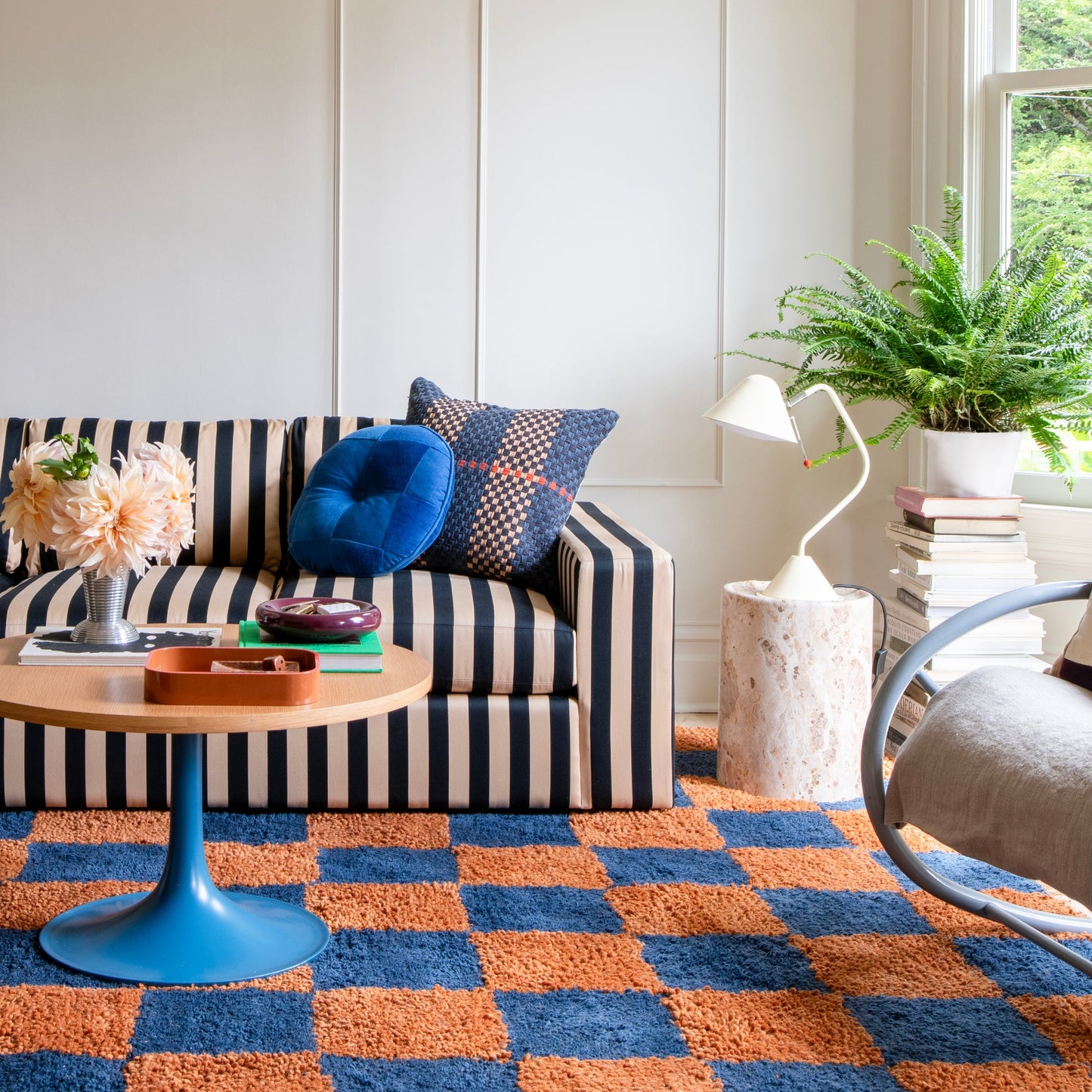 Sunlit living room with a black-and-white striped sofa, blue round pillow, blue-and-orange checkered rug, round wooden coffee table, and white task lamp on a stone side table