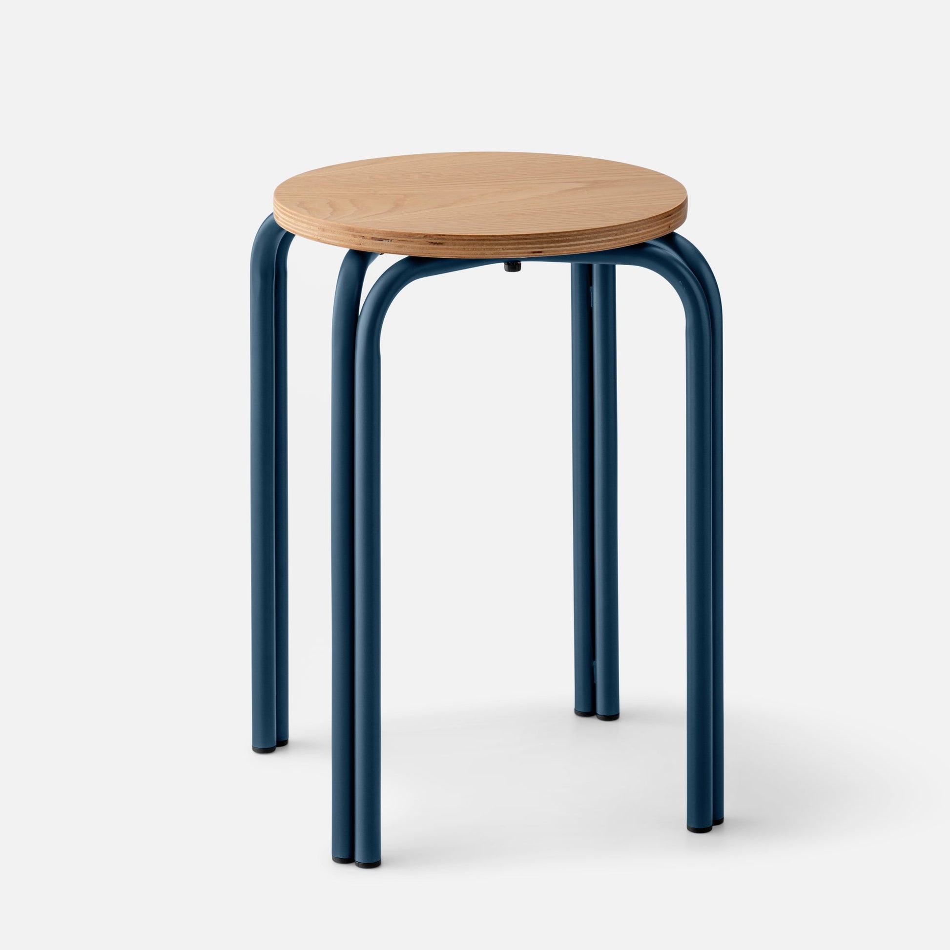 Three-quarter view of Forma Stool showing round white oak top with exposed plywood edge and indigo powder-coated steel legs on light gray background