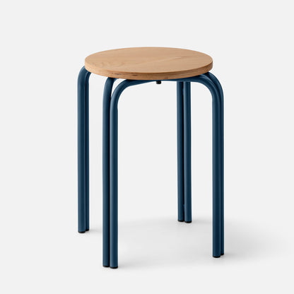 Three-quarter view of Forma Stool showing round white oak top with exposed plywood edge and indigo powder-coated steel legs on light gray background