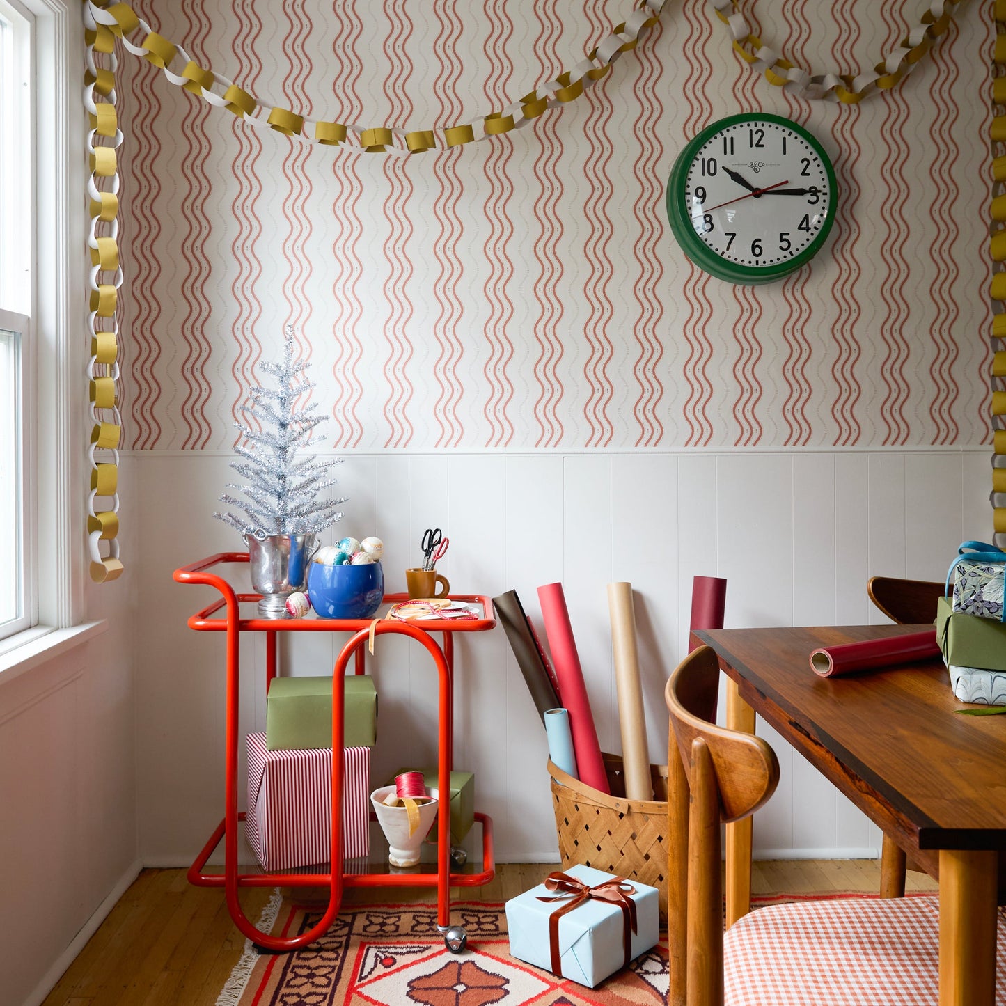 Three-quarter view of red Lolly Bar Cart with bent-steel frame, two glass shelves, and caster wheels in a gift-wrapping corner
