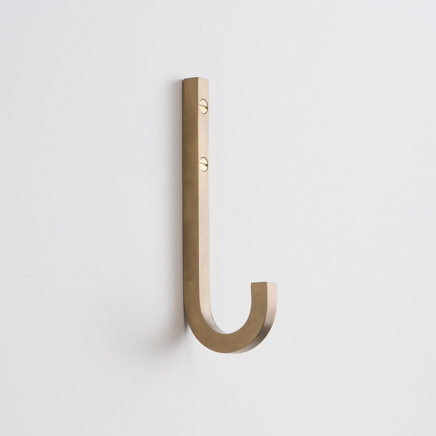 Angled view of J Hook in natural brass showing square solid-brass profile and two visible mounting screws on white wall