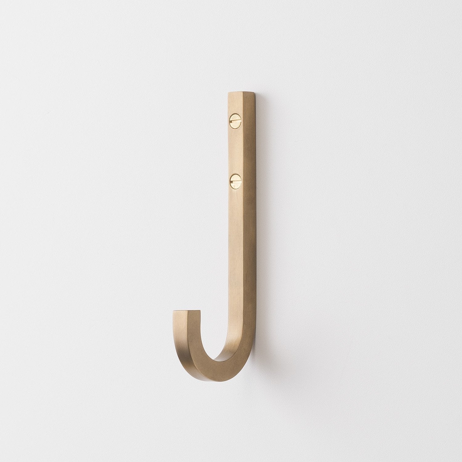 Front view of J Hook in natural brass showcasing solid brass finish, visible mounting screws, and curved J shape on a white wall