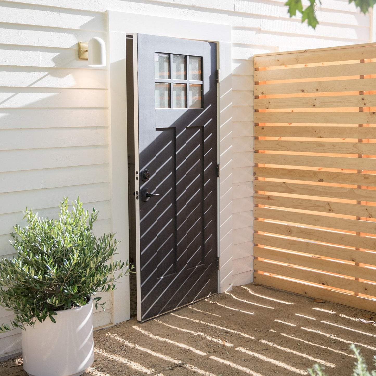 White dome wall sconce mounted beside a black entry door on a sunlit exterior with a wooden slat privacy screen