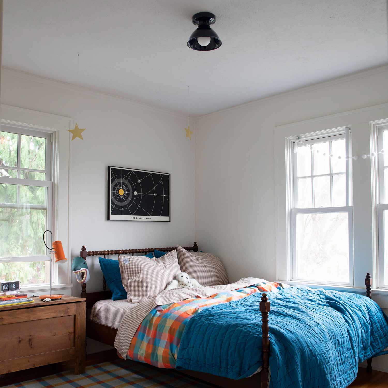 Gingham pillowcases on a styled bed in a bright corner bedroom with a blue quilt, wooden nightstand, and two windows