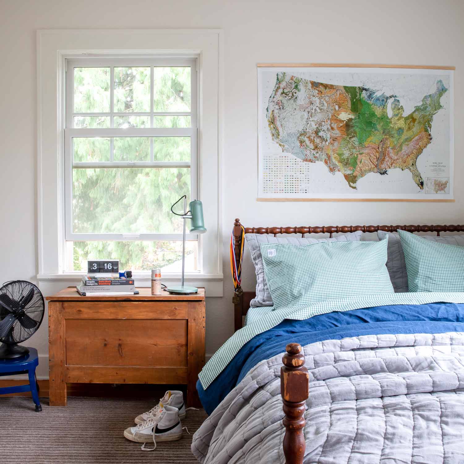 Two juniper green gingham pillowcases on a bed with blue gray bedding, a wooden nightstand and a wall map