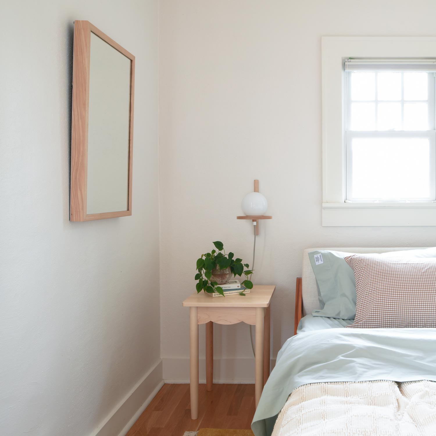 Gingham pillowcases in juniper green on a made bed beside a wooden nightstand with wall sconce, mirror, and potted plant
