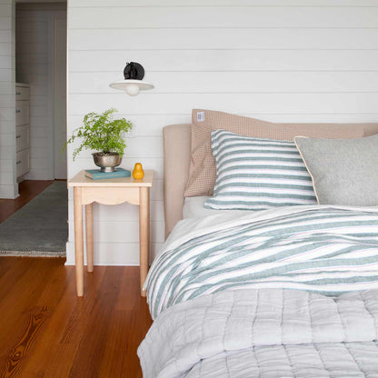 Bed styled with gingham pillowcases, striped and gray accent pillows, and a nightstand with a plant and wall sconce