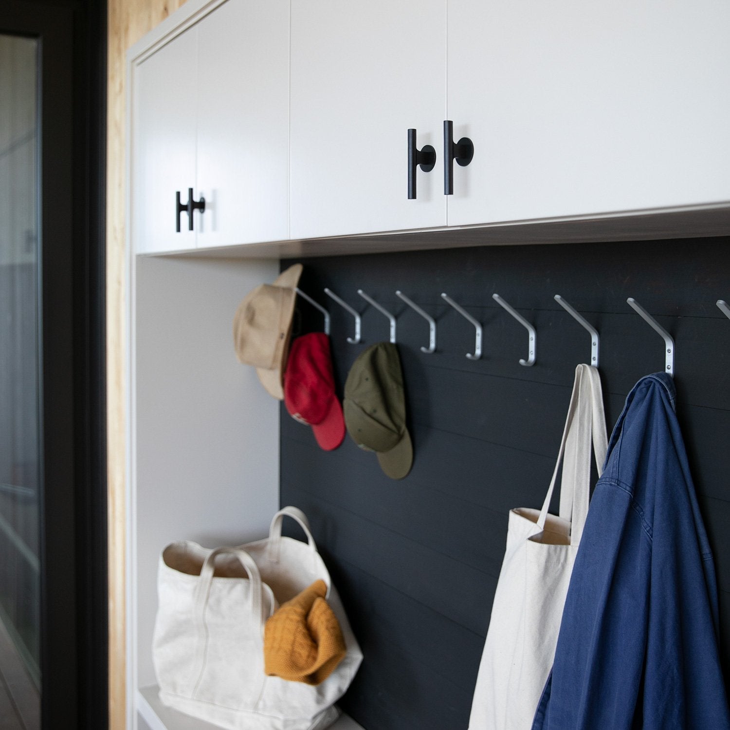 Two black Galley Pulls mounted on white upper cabinets above a mudroom bench with hooks holding hats, tote bags, and a blue jacket