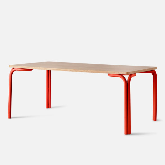 Angled side view of Forma Dining Table showing light wood top with rounded corners and red tubular legs on white