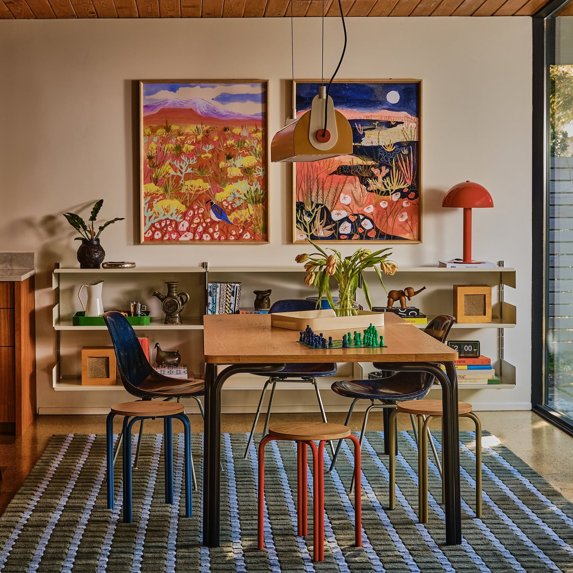 Oak top stools with colored metal legs around a dining table with two framed prints and a pendant lamp