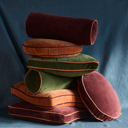 Angled side view of Velvet Piped Pillow stack, velvet with contrast piping in raisin, rust, green on blue backdrop